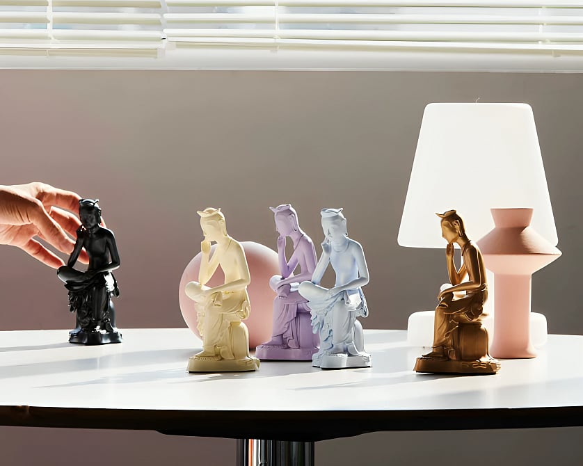 A bright, minimalist studio photograph of five small, identical pensive Bodhisattva statues arranged in a row on a white round table. Each statue is a different solid color: black, cream, lavender, pale blue, and metallic gold. On the far left, a person's hand reaches out toward the black statue. In the background, a soft pink sphere sits behind the central figures, while a glowing white table lamp and a pink geometric vase stand on the right. Sunlight filters through horizontal window blinds from above, casting soft shadows across the scene.
