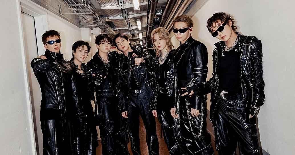A group photograph of seven young men posing closely together in a long, narrow hallway with white walls and an industrial ceiling featuring exposed pipes and light fixtures. They are dressed in coordinated, edgy black leather outfits, including jackets and pants heavily detailed with silver studs, zippers, and draped chains. Three of the men are wearing dark sunglasses. They face forward toward the camera with confident expressions under bright, overhead hallway lighting. BTS members RM (알엠, Kim Namjoon/김남준), Jin (진, Kim Seokjin/김석진), SUGA (슈가, Min Yoongi/민윤기), j-hope (제이홉, Jung Hoseok/정호석), Jimin (지민, Park Jimin/박지민), V (뷔, Kim Taehyung/김태형), and Jung Kook (정국, Jeon Jungkook/전정국). BTS in Brussels. BTS EUROPE TOUR 2026. ARIRANG.