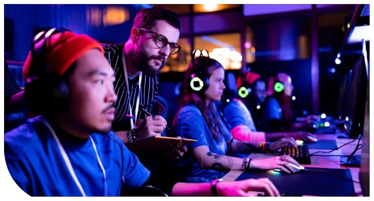 A mentor stands behind gaming students wearing headsets and playing computers in a room lit with blue and purple neon lights.