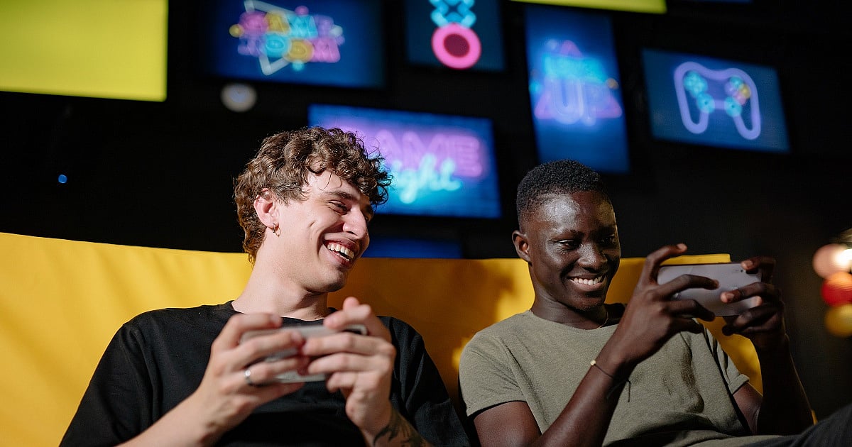 Two people sit side by side on a yellow couch, smiling while playing games on their smartphones. One wears a black T-shirt and laughs while holding a phone in both hands, while the other wears a light green T-shirt and focuses on the screen with a grin. Neon gaming-themed signs create the atmosphere of a modern gaming lounge.