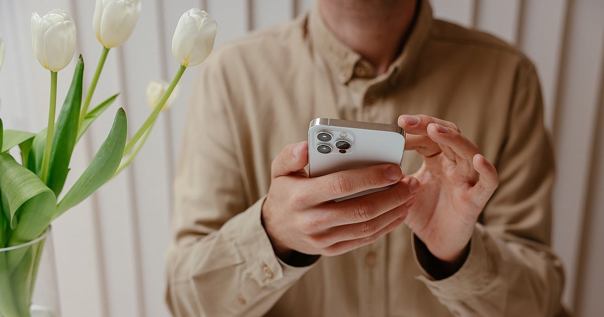 A close-up of a person in a beige button-up shirt using a silver smartphone with one hand while tapping the screen with the other. White tulips in a vase are positioned in the foreground on the left, while soft, neutral curtains create a bright, minimalist indoor background.
