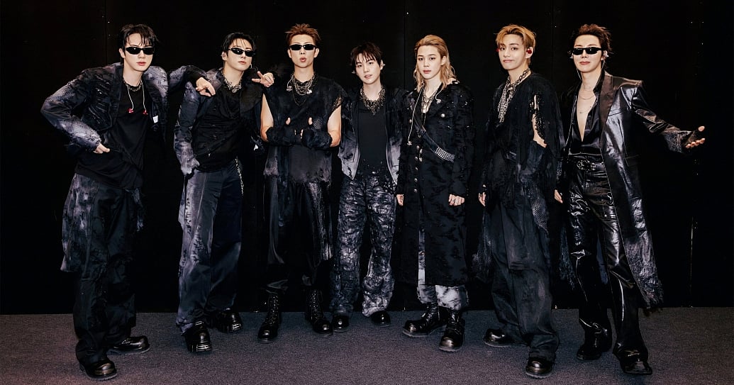 All seven members of K-pop group BTS—Jin, Jung Kook, RM, Suga, Jimin, V, and j-hope—standing side-by-side in coordinated all-black avant-garde streetwear, layered with silver chains and sunglasses, posing against a solid black backdrop.