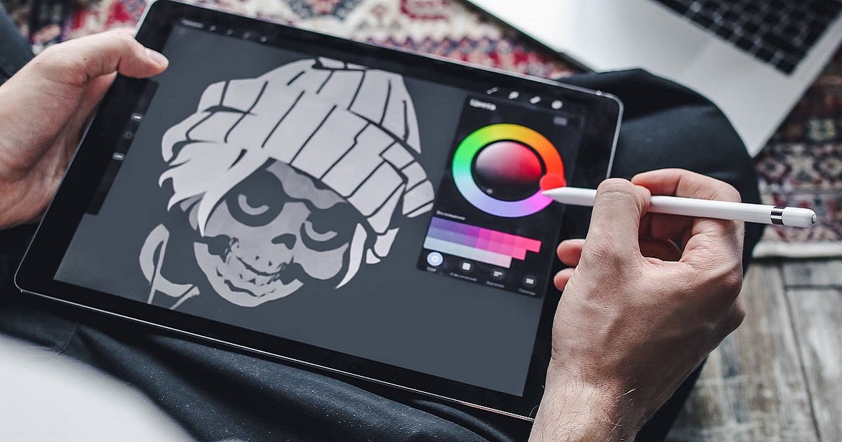 A person uses a white stylus to draw a monochrome cartoon-style skull character wearing a knitted beanie on a tablet. The tablet screen displays a digital art app with a circular color wheel and brush controls on the right, while the artist holds the device with the other hand.