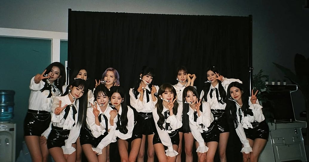 A flash-lit group photo of all 12 members of K-pop group IZ*ONE posing backstage against a dark curtain backdrop. They wear coordinated stage outfits featuring long-sleeved white collared blouses with black neck ribbons or bows, paired with shiny black high-waisted leather shorts or skirts, and thin headset microphones. The members stand close together in staggered rows, smiling and gesturing at the camera with peace signs and hand hearts.