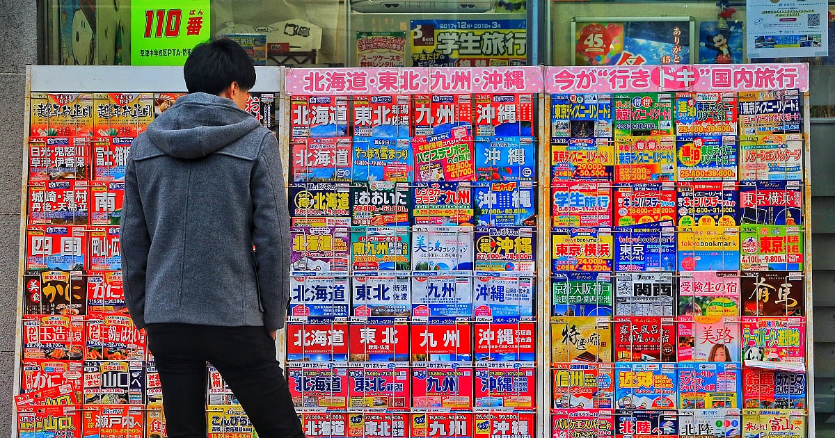 A man in a grey hoodie and black pants browsing a vibrant magazine stand in front of a toy store on a street in Otsu, Japan, showcasing several magazines up for sale.