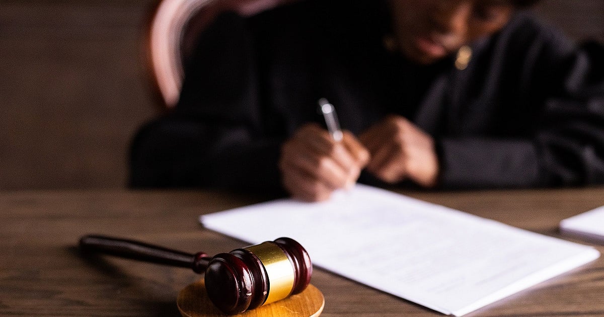 A judge in official uniform signing printed documents at desk in what appears to be a courtroom. The focus of the image is on the gavel, representing law and justice.