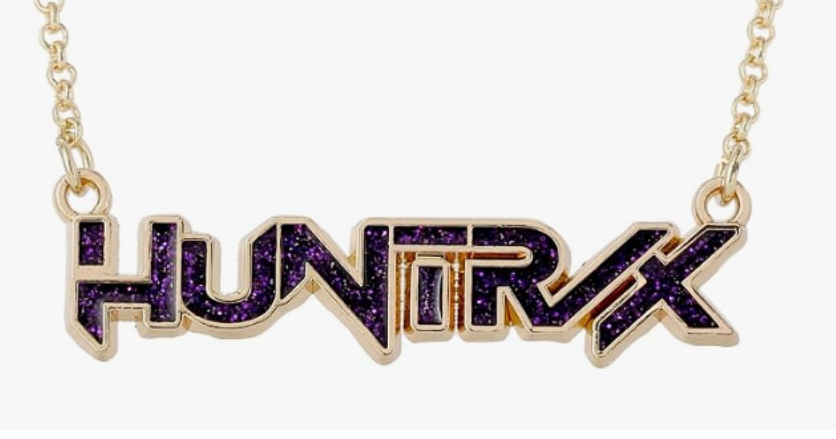 Gold-tone HUNTR/X nameplate necklace with purple glitter detailing on a white background.