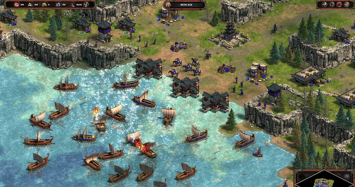 A Age of Empires II: Definitive Edition screenshot showing a civilization battling across a river with warships, cavalry, buildings, and fortified cliffs.