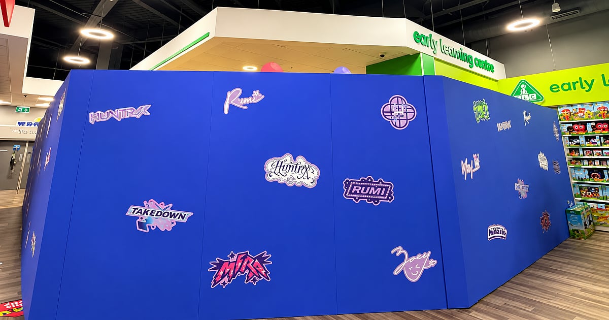 Blue KPop Demon Hunters-branded retail installation featuring HUNTR/X logos and character decals on temporary partition walls inside a shopping centre, with toy displays and the Early Learning Centre sign in the background.