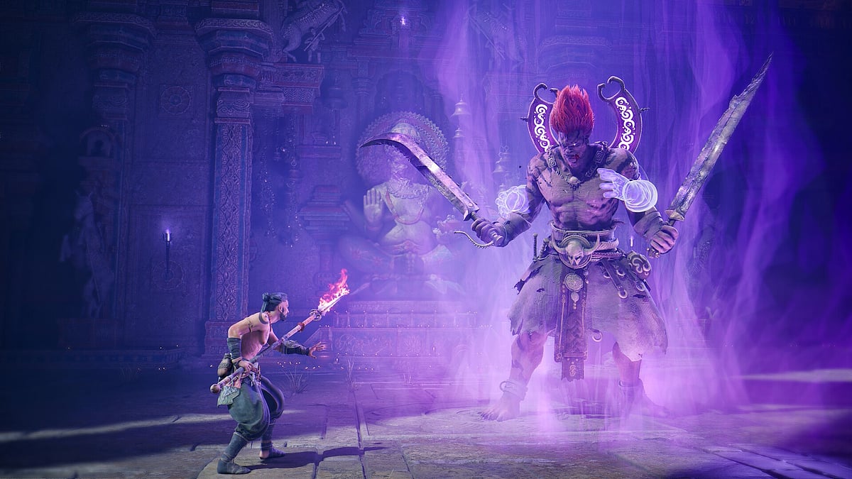 A screenshot from a gameplay trailer of Age of Bhaarat. The gameplay segment shows the protagonist wielding a flaming spear facing off against a giant boss in a stone-walled arena who is staring him down dual wielding two weapons.