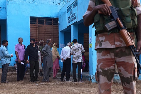J&K Assembly elections, 2nd Phase voting: A security personnel stands guard as people wait to cast their votes in Rajouri district