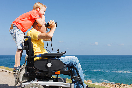 A father in a motorized wheelchair carries a smiling boy on his shoulders.