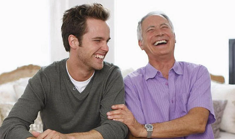 An Old Man And Young Man Sitting Together on a couch, laughing and enjoying each others company.