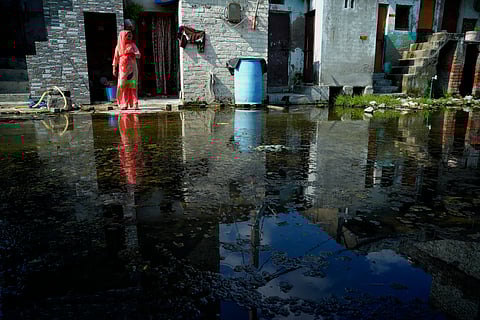 A woman stands at the entrance of her home, surrounded by stagnant wastewater flooding the street after rains. 