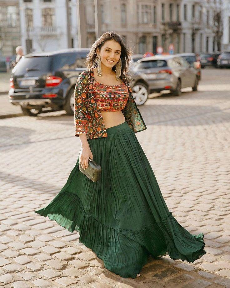 A woman in a dark teal lehenga and a patterned choli, smiling outdoors.