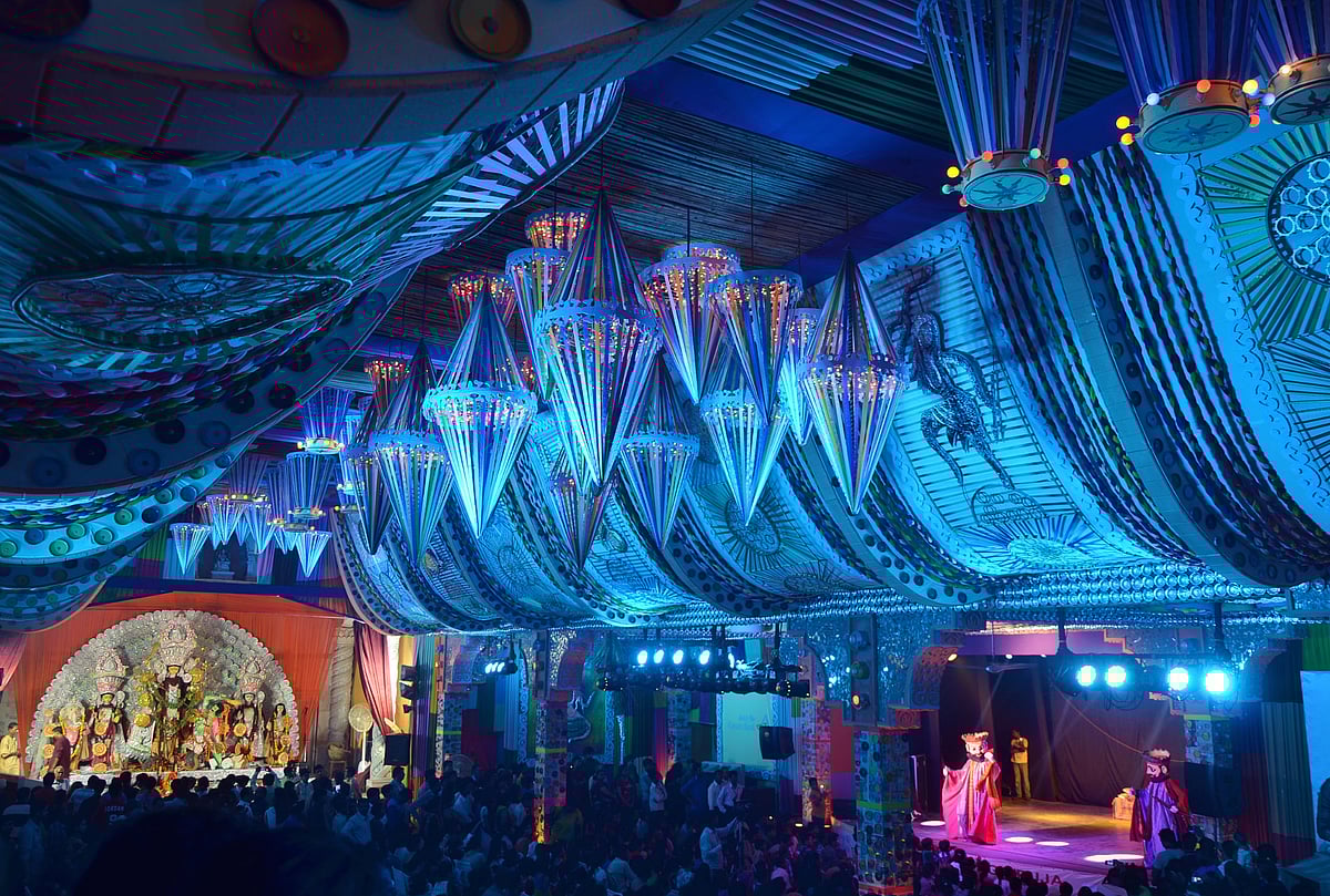 An ornate pandal with a blue draped ceiling and a stage with performers and an idol.