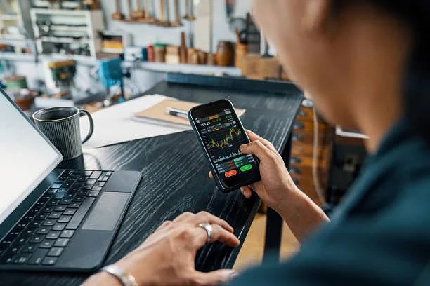 Person checking stock market charts and trading app on smartphone while working at desk with laptop