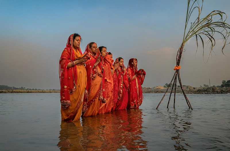 Women in orange and yellow sarees standing in water for the Chhath Puja ritual.
