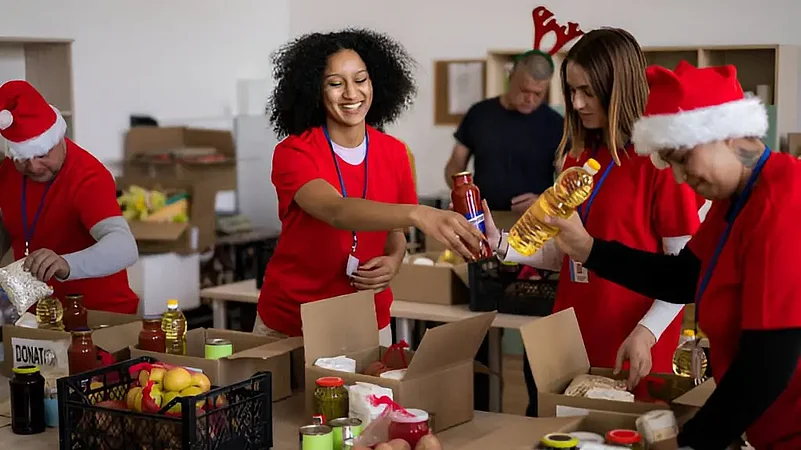 Smiling people wearing red shirts and Santa hats pack food donations at a Charity Corner.
