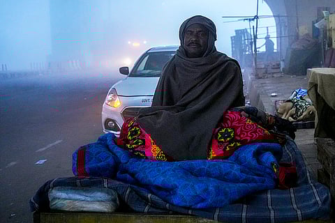 A man wearing a shawl rests on a roadside amid dense fog on a winter morning, at Mayur Vihar area, in New Delhi. Visibility plunged across parts of Delhi on Saturday morning, with buildings and flyovers fading into a grey blur amid dense fog, as the capital’s AQI edged closer to the ‘severe’  category at 384. 
