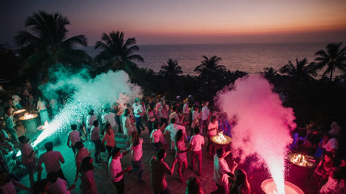 Crowded outdoor party at dusk with blue and pink smoke effects and an ocean backdrop.