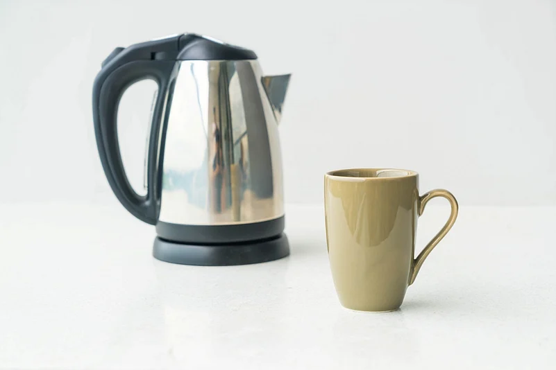 Stainless steel electric kettle with a black handle next to a beige ceramic mug.