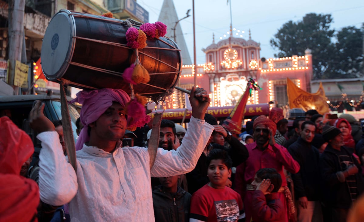 A man in traditional attire carries a large dhol drum on his shoulder during a festive parade.