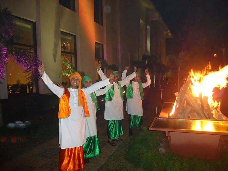 Performers in traditional attire dancing with arms raised near a large outdoor bonfire at night.