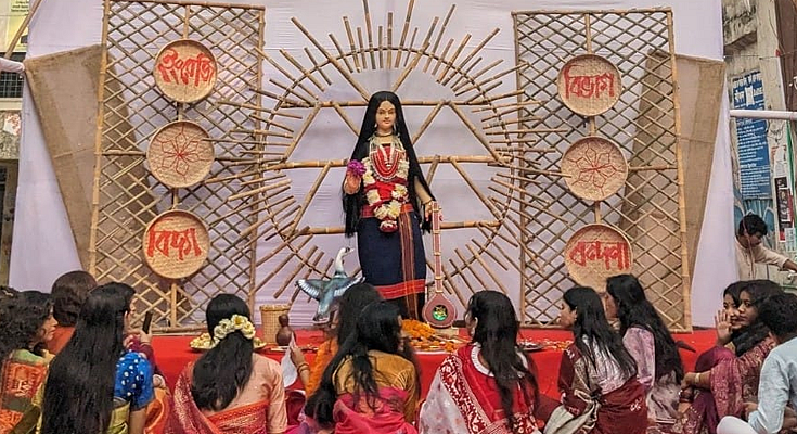 An idol of Goddess Saraswati on a stage with devotees gathered for a puja ceremony