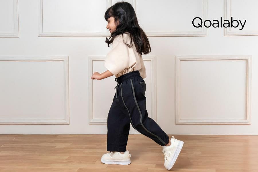 A young girl in black pants, a beige shirt, and white sneakers walks; Qoalaby logo visible