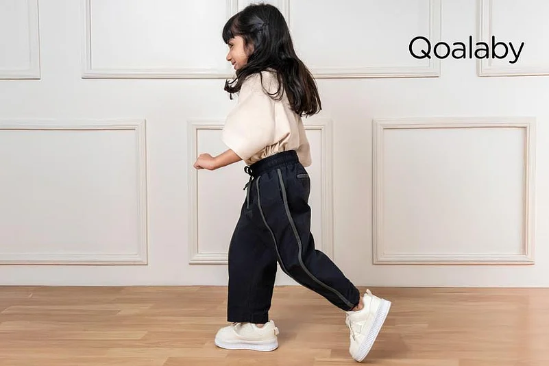 A young girl in black pants, a beige shirt, and white sneakers walks; Qoalaby logo visible