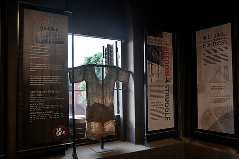 The image displays an exhibit at the Cellular Jail, The central item is a replica of the coarse jute uniform worn by prisoners during their incarceration for involvement in India's freedom struggle.