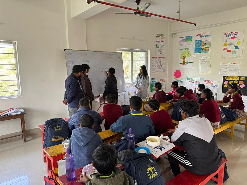 A teacher instructs students in a red-desk classroom setting
