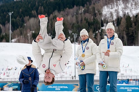 Silver medalist Ben Ogden, of the United States, does a back flip from the podium while gold medalist Johannes Hoesflot Klaebo, of Norway, and bronze medalist Oskar Opstad Vike, also of Norway, right, look on after the cross-country skiing men's sprint classic at the 2026 Winter Olympics, in Tesero, Italy.