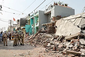 | Photo: PTI : Police personnel stands guard as a demolition drive is underway to clear illegal houses along the Aji river banks and TP Road in the Jangleshwar area, in Rajkot, Gujarat.