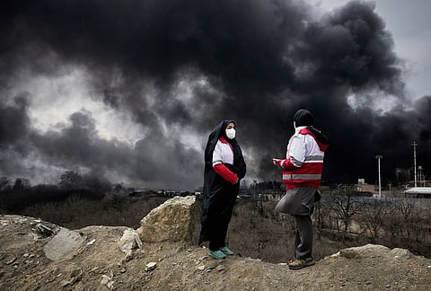 Two women from the Iranian Red Crescent Society stand as a thick plume of smoke from a U.S.-Israeli strike on an oil storage facility late Saturday rises in the sky in Tehran, Iran, Sunday, March 8, 2026. 