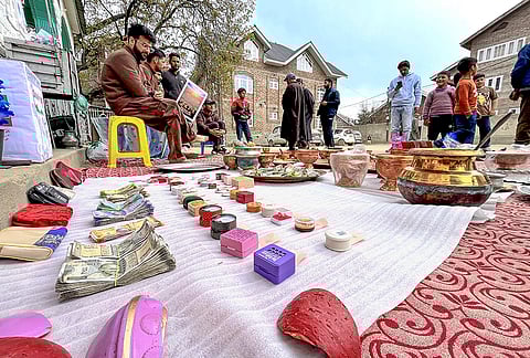 People sit near items including cash, gold, silverware and copper utensils received as contribution by the public during a donation drive for the war-ravaged Iran, at Delina, in Baramulla district, Jammu and Kashmir.