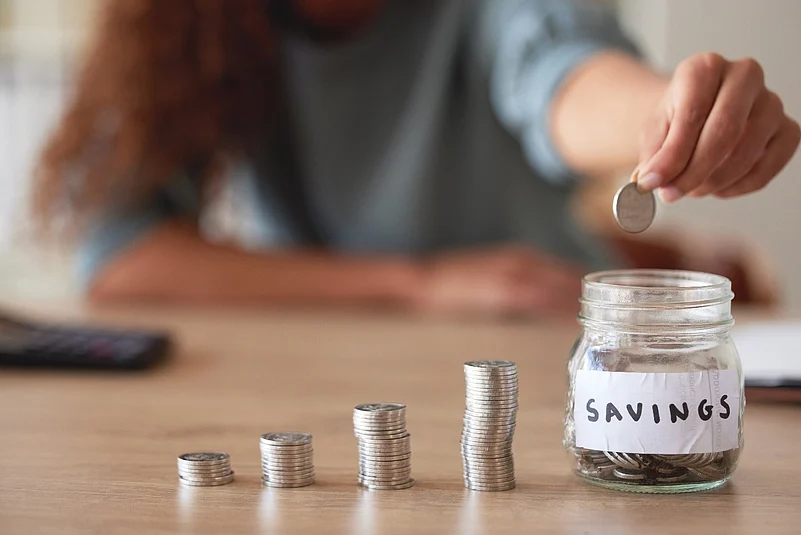 Person dropping a coin into a labeled savings jar beside growing stacks of coins