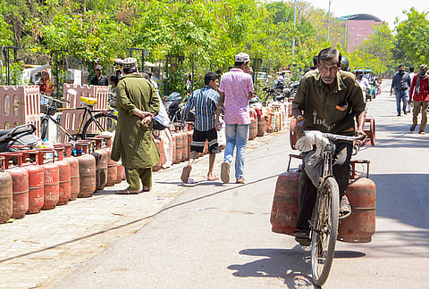 A gas agency worker carrying LPG cylinders on a bicycle rides past people sitting near empty cylinders placed in a queue outside a gas agency, amid disruptions in LPG supplies due to the ongoing West Asia war, in Kanpur, Uttar Pradesh.