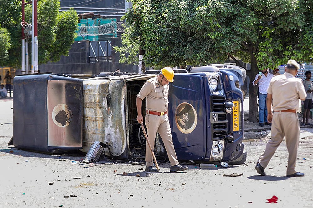 Police personnel inspect the wreckage of a vehicle during a protest by factory workers demanding a hike in wages, in Noida, Gautam Buddh Nagar district, Uttar Pradesh. The protest carried incidents of arson, vandalism and stone-pelting reported from Phase-2 and Sector 60 areas, police said.  - | Photo: PTI