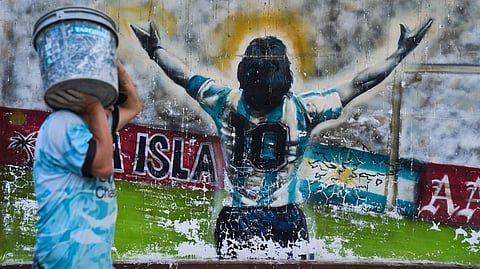 A pedestrian carries a bucket past graffiti depicting late soccer legend Diego Maradona in Buenos Aires, Argentina, Monday, April 13, 2026