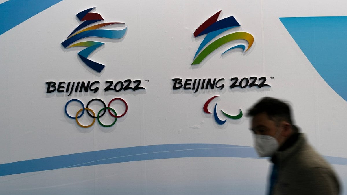 A man wearing a KN95 mask walks past the logos of the 2022 Winter Olympics and Paralympics.