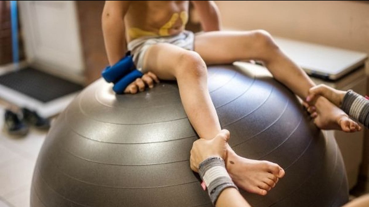 Physical therapist assisting little boy make exercises on gym ball during rehabilitation