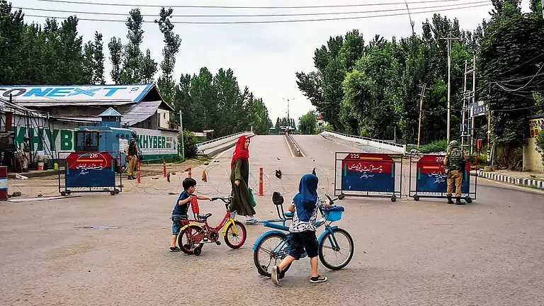 The Kashmir Life: A woman walks with her children as security personnel stand guard - null