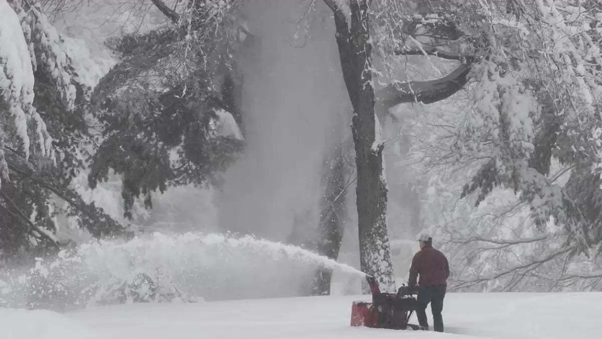 A man uses a snow blower to make a pathway in his yard, as snow falls around him. 