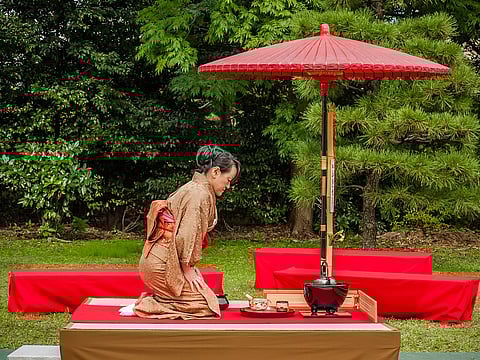 Tea being prepared outdoors called nodate, underneath a red parasol called nodatekasa. The hostess wears a brown kimono.
