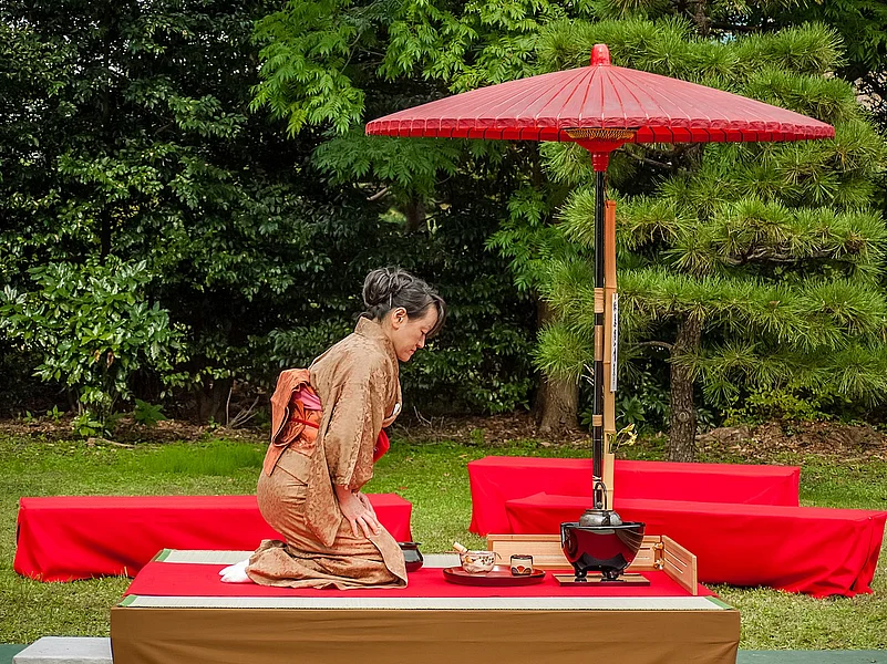 Tea being prepared outdoors called nodate, underneath a red parasol called nodatekasa. The hostess wears a brown kimono.