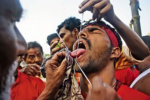 A priest deftly pierces a Gajan sanyasi’s tongue with a metal needle, drawing little blood—his skill ensures minimal harm during this deeply symbolic ritual act