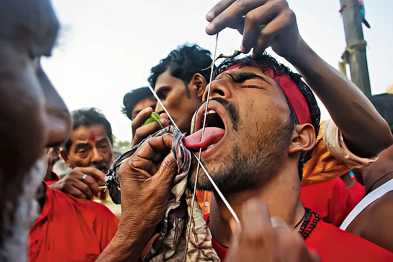 A priest deftly pierces a Gajan sanyasi’s tongue with a metal needle, drawing little blood—his skill ensures minimal harm during this deeply symbolic ritual act