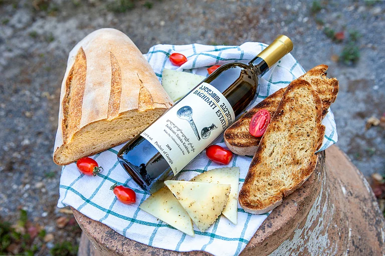 A bottle of Georgian natural wine with fresh bread, cheese, and cherry tomatoes - Photo by ALEXANDRA TORRO on Unsplash